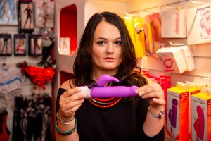 Lucy stands in LUST sex shop in Brighton holding a pink rabbit vibe with a small lil pout, in front of the toys