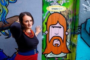 Lucy stands with her hand behind her head next to a graffiti picture of Jesus, with red hair and a halo. She wears a black top, orange necklace and red trousers. Purity culture. Low libido in women.
