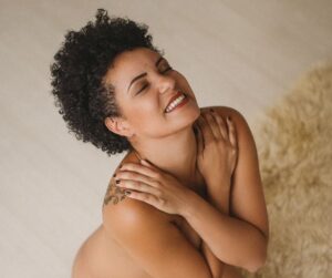 Mixed race woman is naked, kneels on the floor smiling, with her arms crossed over her chesrt. It is a creamy white floor