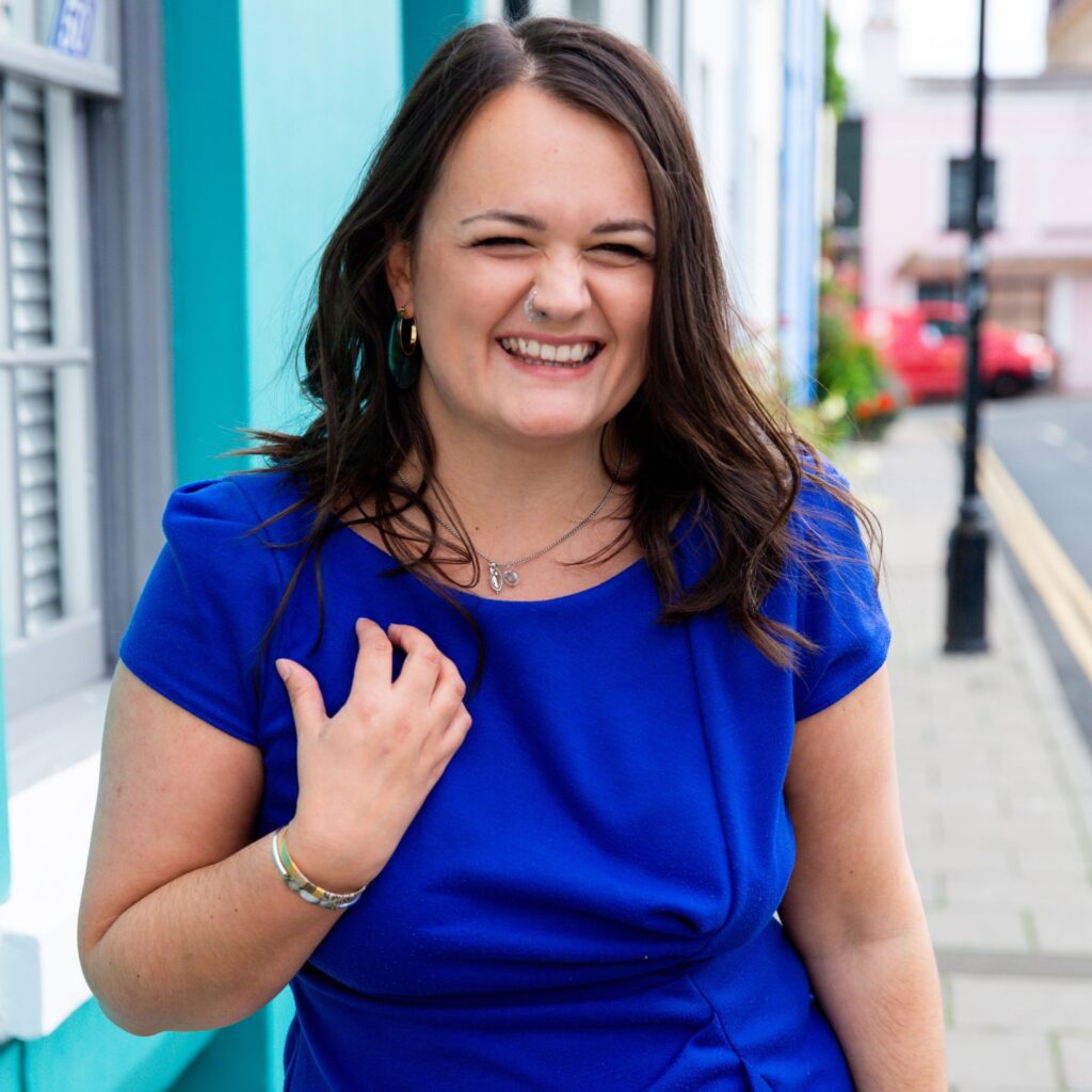 Lucy stands smiling with her hand touching her chest, wearing a blue dress, standing next to a light blue house in Brighton