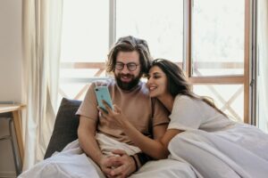 White heterosexual couple sitting in a bed smiling, the woman is holding a phone to the man. The man has a beard and glasses.