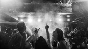 Black and white scene of a gig or a concert, someone with long hair has their arms raised pointing at the ceiling, there is smoke