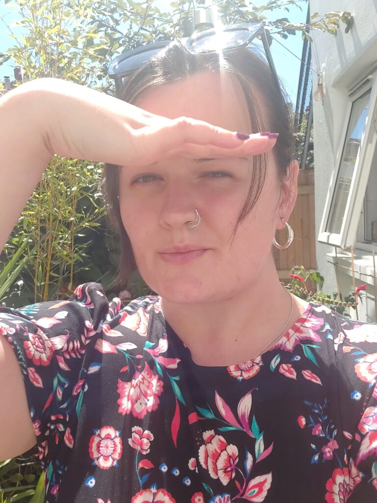 Lucy wears a black and pink dress, with her hand slightly shading her eyes in the garden, with bright sunlight above