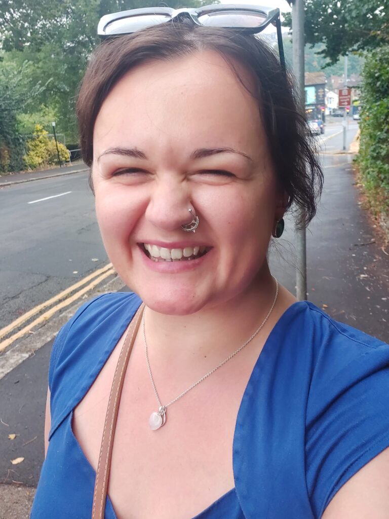 Lucy smiles facing the camera, she wears sunglasses on her head and wears a blue dress. A Brighton street is in the background.